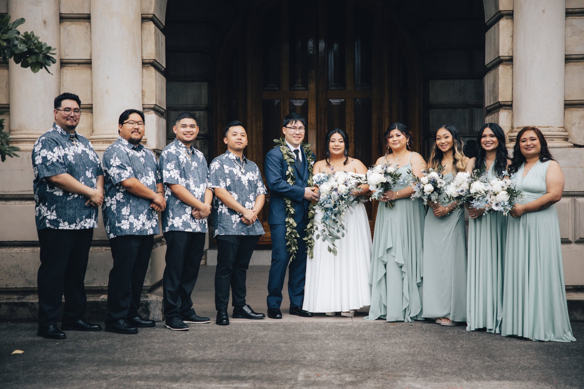 Full wedding party portrait with groomsmen in aloha shirts and bridesmaids in sage green at historic Honolulu venue